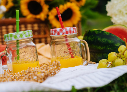 A couple of nice juicr drinks in glass mugs with straws sticking out and fruit beside them, they are all on a picnic sheet
